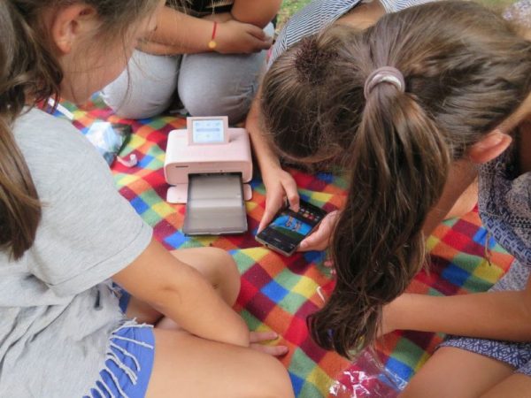 Mehrere Mädchen sitzen auf einer bunten Decke. Alle schauen auf ein Smartphone auf dem eines der Mädchen ein Foto bearbeitet. Daneben steht ein pinker mobiler Fotodrucker.