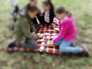 Vier Kinder sitzen auf einer bunten Decke auf einer Wiese und spielen ein Kartenspiel. Das Bild wird zu den Seiten hin sehr unscharf, so das die Kinder nicht zu erkennen sind.