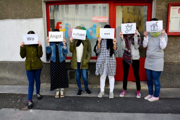 Sechs junge Frauen stehen auf dem Gehsteig. Jede hält ein Papier vor dem Gesicht auf dem entweder ein Wort oder ein Symbol zu sehen ist. Aus den Bildern bildet sich der Satz: "Wo Angst aufhört ist Frieden zuhause."