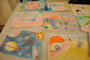 Auf einem Tisch liegen acht bunte Zeichnungen, auf denen jeweils bunte Planeten, UFOs und Aliens gemalt wurden.