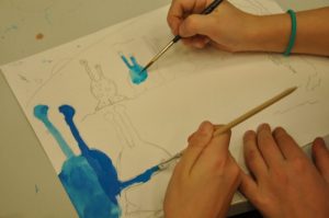 Das Märchen vom Alien ohne zu Hause: Man sieht mehrere Kinderhände, die auf einem weißen Blatt Papier mit blauer Wassermalfarbe Aliens auf einer mit Bleistift vorgezeichneten Planetenlandschaft bemalen.