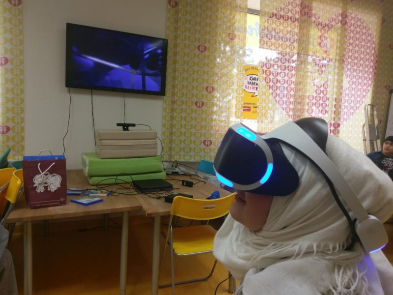 eine junge Frau mit Kopftuch sitzt in einem leicht verdunkelten Raum und hat eine blau leuchtende VR-Brille auf. Im Hintergrund ist ein Fernseher und eine Playstation zu sehen.