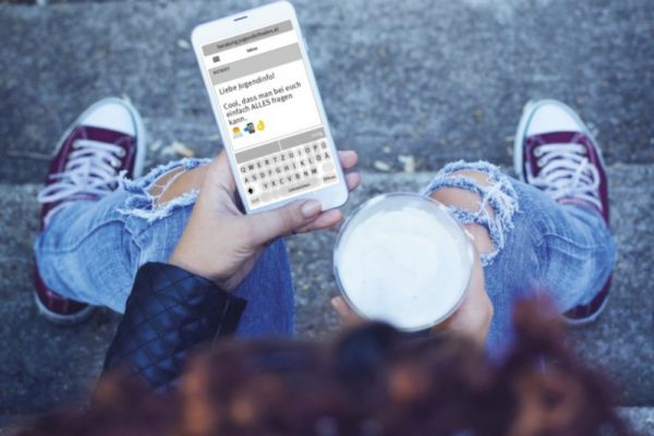 Foto aus der Vogelperspektive: Jemand sitzt auf einer Stufe, hält ein Getränk in der Hand uns schaut auf ein Handy, auf dem ein SMS, das an die Jugendinfo geschickt wurde, zu sehen ist.