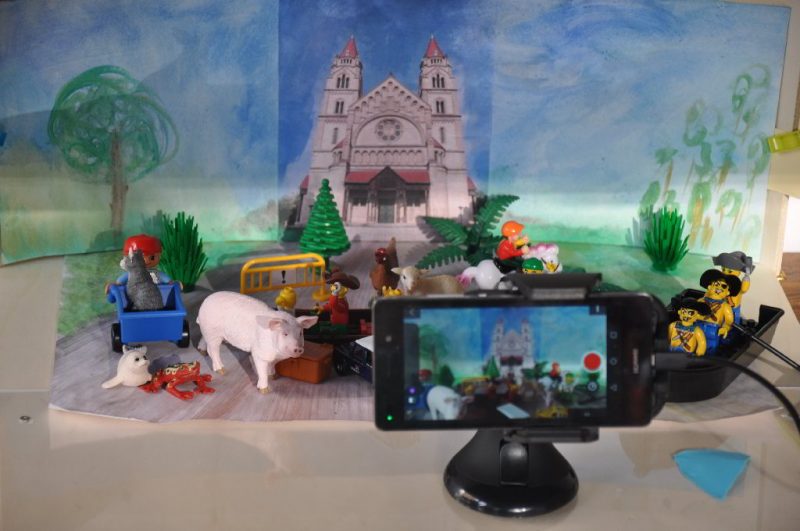 Foto von einer Stopmotion Szene: Verschiedene Plastiktiere und Playmobilfiguren in einem Boot sind vor einer gemalten Kulisse mit einer Kirche und Bäumen zu sehen. Im Vordergrund ein mitfilmendes oder fotografierendes Handy.