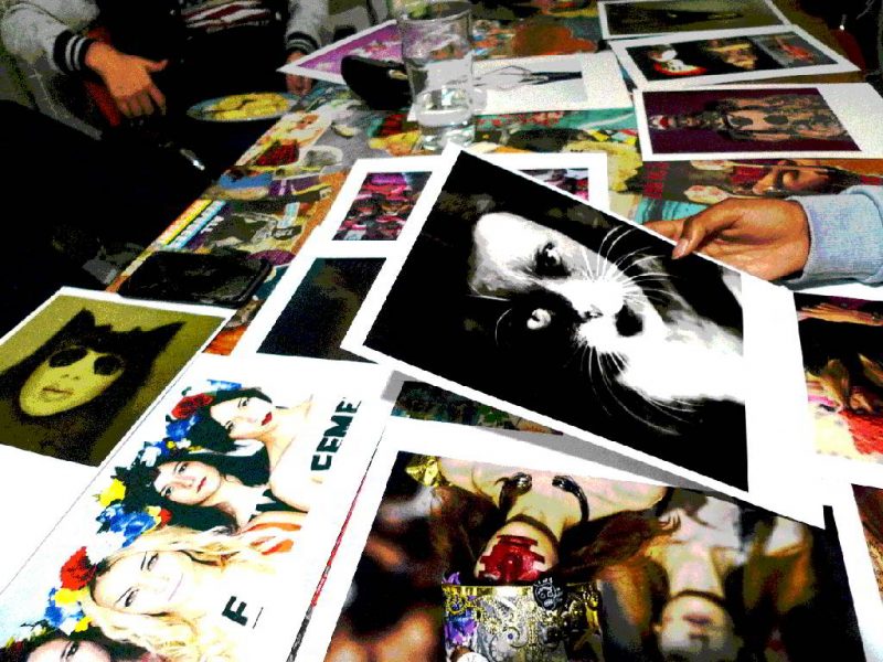 Ausdrucke von verschiedenen Fotos liegen auf einem Tisch, eine Hand hält eine Foto, auf dem ein Gesicht zu sehen ist, welches als Katze verfremdet wurde.
