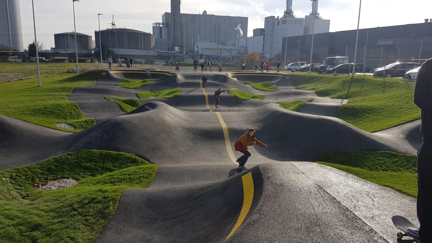 Drei Jugendliche fahren mit ihren Skateboards auf einer Pumptrack, im Hintergrund sind zusehende Menschen zu sehen und eine Industrieanlage in Oberösterreich.