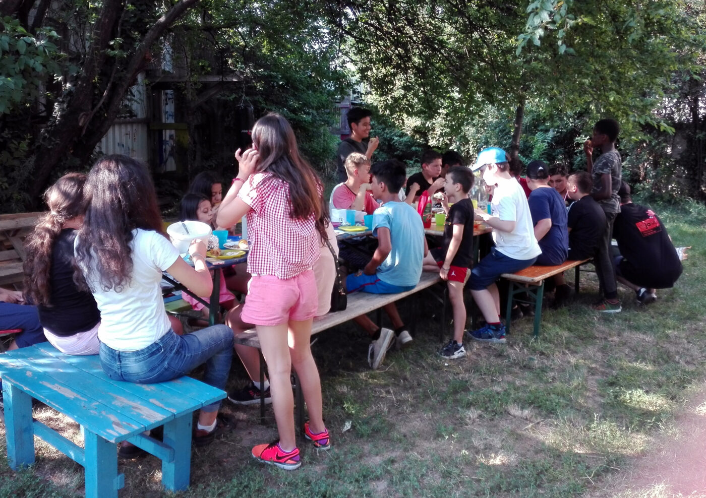 Ca 15 Kinder sitzen auf Holzbänken rund um mehrere Tische und essen und trinken und unterhalten sich.