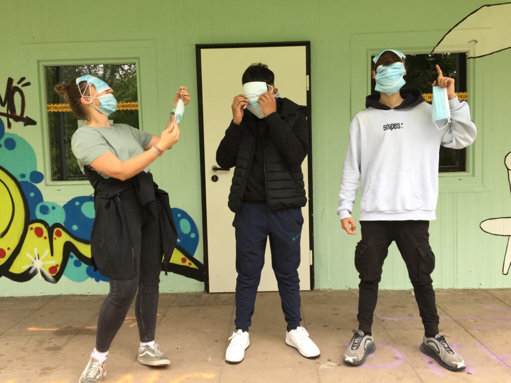 Ein launiges Foto zeigt drei junge Menschen, die mehrere Mund-Nasen-Schutz-Masken auf dem Kopf und im Gesicht aufhaben. Sie stehen vor einer Wand mit Türe und Fenstern. Die Wand ist grün und hat ein Graffiti.