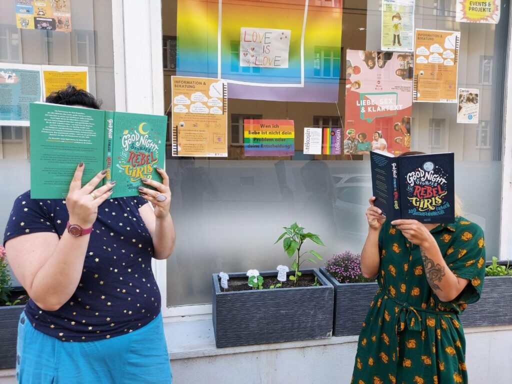 Zwei Frauen in bunten Klamotten stehen vor einem Auslagenfenster. Sie stehen und lesen intensiv das Buch "Good Night Stories for Rebel Girls: 100 außergewöhnliche Frauen."