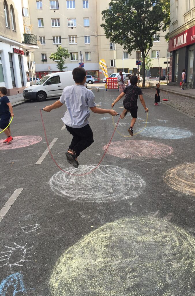 Kinder spielen Seilhüpfen auf einer Spielstraße in Wien. Die Straße ist mit bunten Straßenkreidekreisen bemalt. Es wird auf genügend Abstand auf Grund der COVID19 Pandemie geachtet.