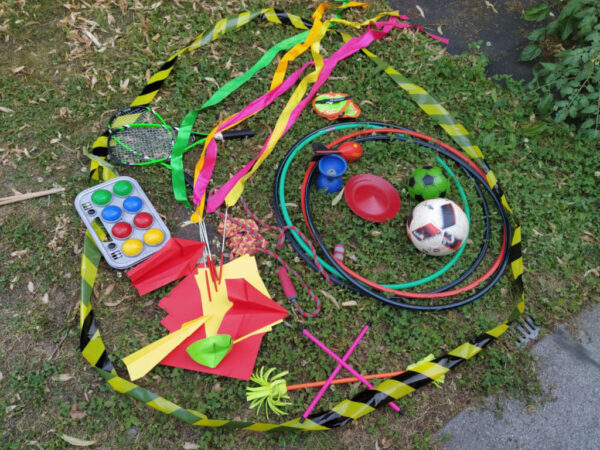 Das Foto zeigt buntes Spielmaterial auf einer Wiese liegend, wie zum Beispiel HoolaHoop Reifen, Boggia Kugeln, Badminton Schläger, Pois, Fußbälle, Diabolo Sticks uvm.