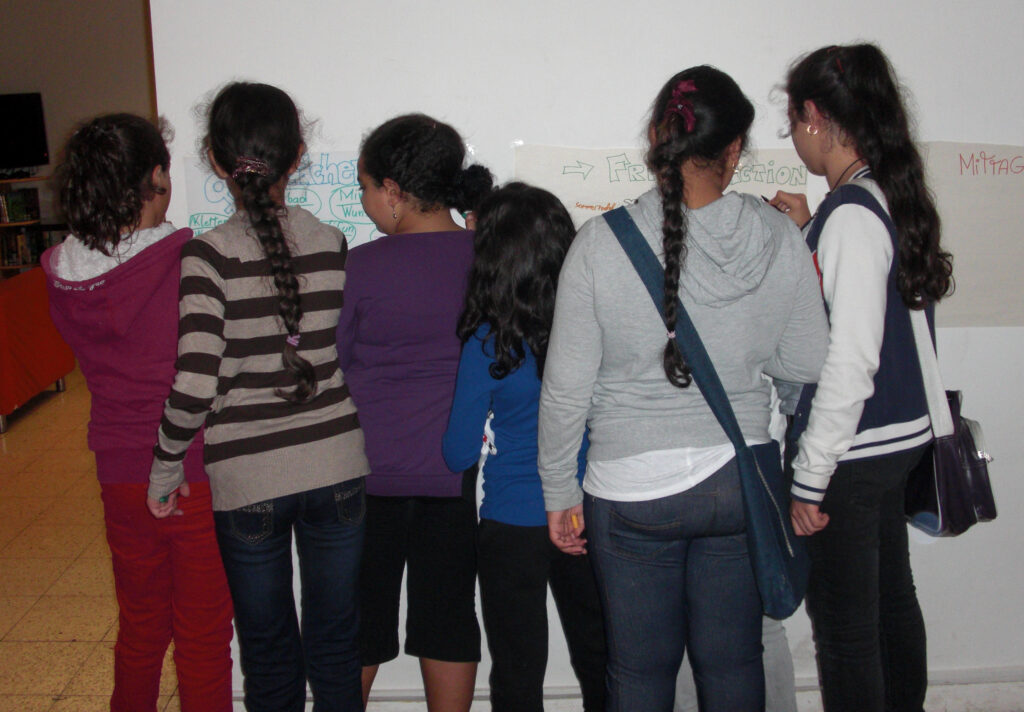 Eine Gruppe von Mädchen ist von hinten zu sehen, wie sie etwas auf Plakate an der Wand eintragen