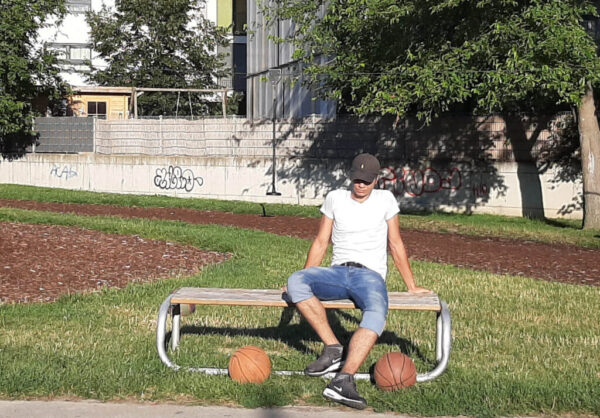 Ein junger Erwachsener sitzt auf einer Bank in einem Park. Er trägt eine braune Kappe, weißes T-Shirt und eine kurze Blue-Jeans und Blickt zu Boden. Vor ihm liegen zwei Basketbälle.