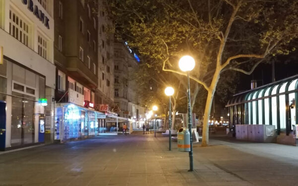 Eine menschenleere Straße bei Dunkelheit, in der Mitte ist eine Reihe von Straßenlaternen. Am linken Bildrand sind Auslagen von Geschäftslokale zu sehen. Am rechten Bildrand ist eine U-Bahn-Station zu erkennen.