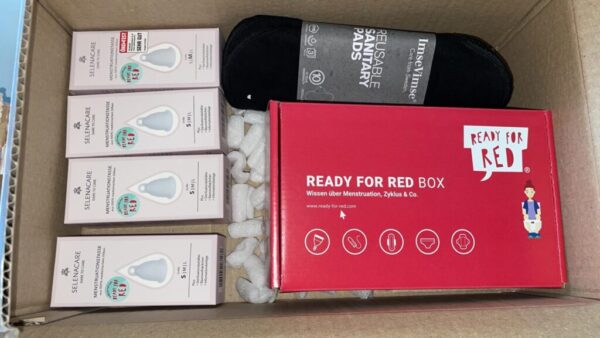 Ein Karton, der mit vier Menstruationstassen, einer Packung wiederverwendbaren Binden und einer Schachtel mit Infomaterial zum Thema Menstruation gefüllt ist.