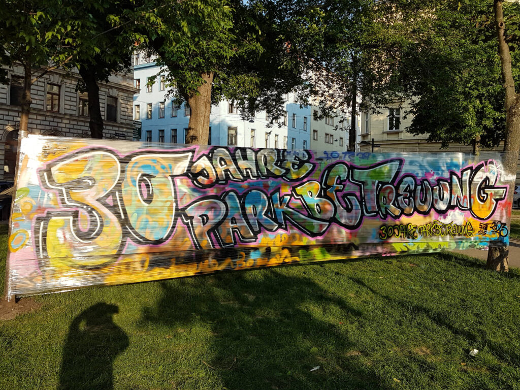 Auf einem Banner, das zwischen zwei Bäumen aufgespannt wurde, steht „30 Jahre Parkbetreuung“ in Graffiti Schrift geschrieben.